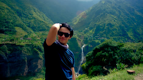 young man in sunglasses points toward camera in green hilly nature