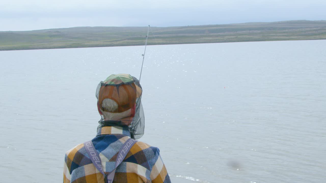 Fisherman Wearing A Cap And Mosquito Netting Sits By A Lake With Fishing Rod In Hand. - closeup shot