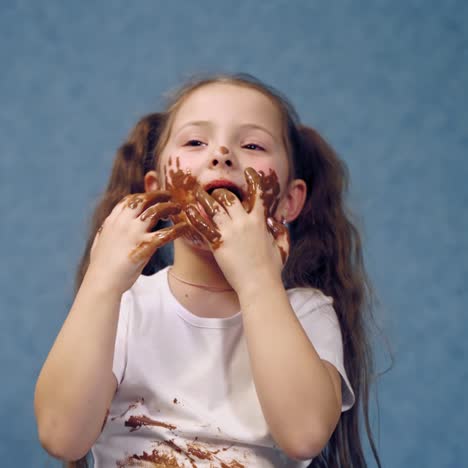 smiling child licking chocolate off her fingers. funny little girl with two ponytails has dirty hands and face after eating chocolate on blue studio background.