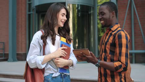 mujer caucásica y hombre afroamericano hablando y viendo algo en la tableta en la calle cerca de la universidad