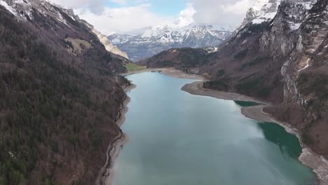 An-aerial-perspective-captures-Klöntalersee-in-Glarus,-Switzerland,-framed-by-the-towering-Vorderglärnisch-mountain-and-embraced-by-snow-capped-peaks,-the-captivating-beauty-of-the-Swiss-Alps