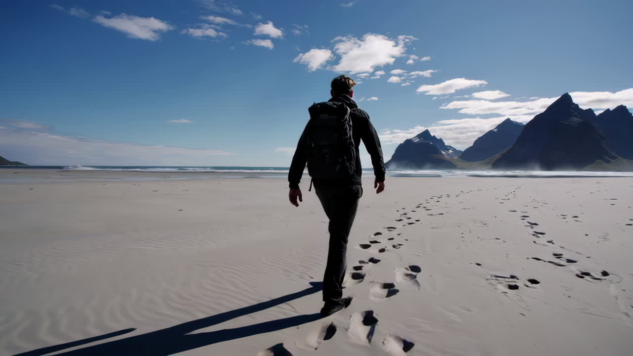 Person with backpack walking on a vast sandy beach with mountains and sea