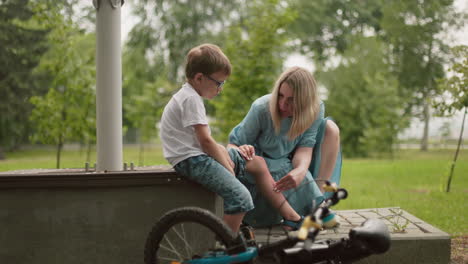 a mother kneels to clean her son's wound while they sit along a tarred road, the boy holds his trouser, looking sad, as his fallen bicycle lies nearby, the mother tenderly wipes his tears