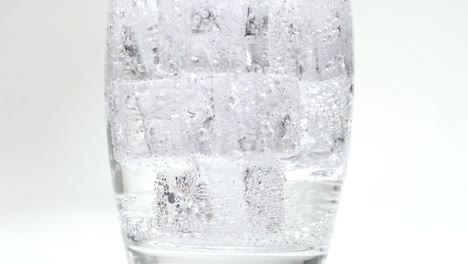 soda or sparkling water pouring onto ice cubes filling up glass in slow motion bubbles close up carbonation white studio background