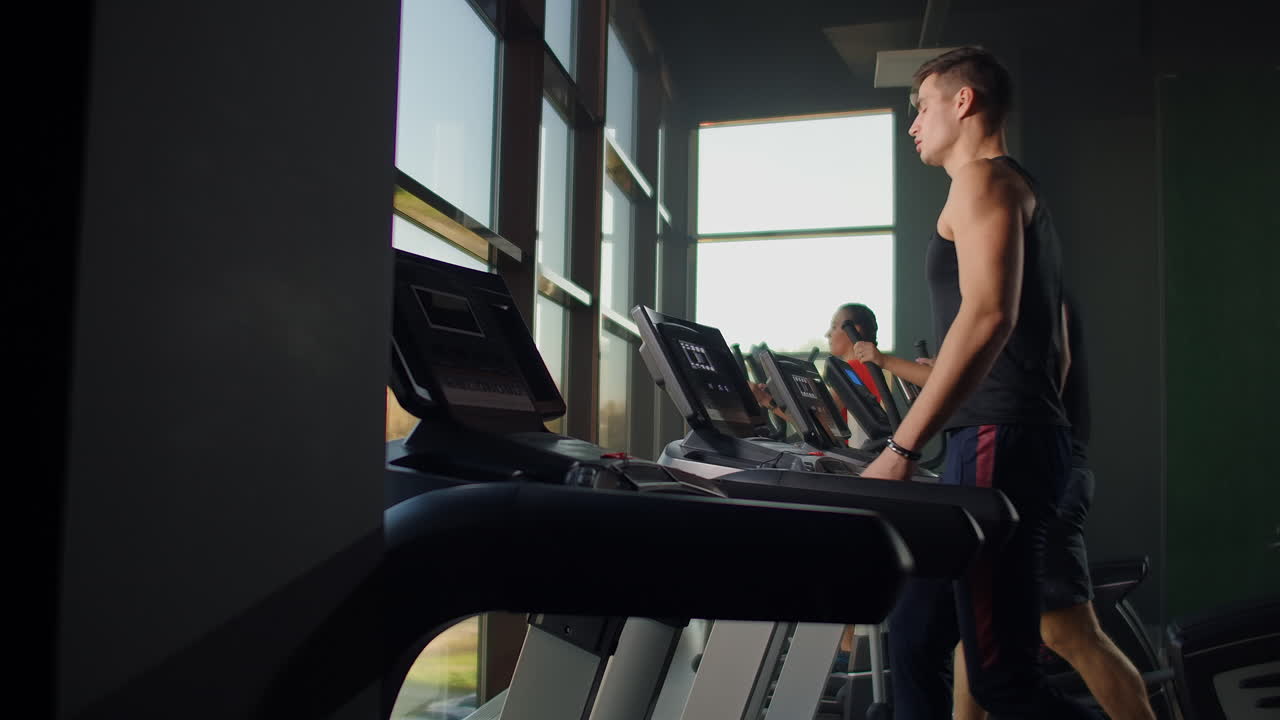 Men walk on treadmills in the fitness room. A group of people walking on treadmills near a large panoramic window. Start of fitness classes. Young beautiful women and men in sportswear in cardio fitness room