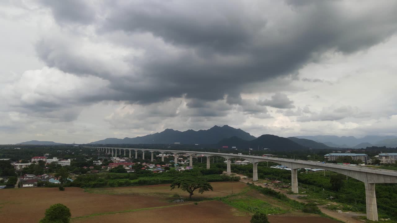4k 공중 하강 샷은 태국 사라부리에 있는 프레임을 가로지르는 미래형 고가 철도로 우레와 같은 우울한 날씨의 무악 렉 산악 실루엣을 보여줍니다.