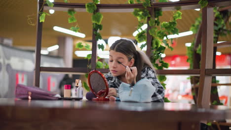 young lady brushing her eyelashes with mascara in front of a mirror, with blurred background of people walking in a public place, creating a vibrant and busy atmosphere in a makeup routine