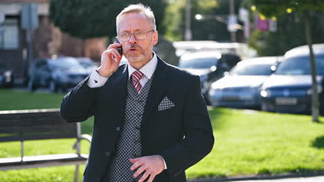 outdoor portrait of mature businessman talking on mobile phone. senior executive businessman with beard and glasses solves the problems while talking on cell phone in the city.