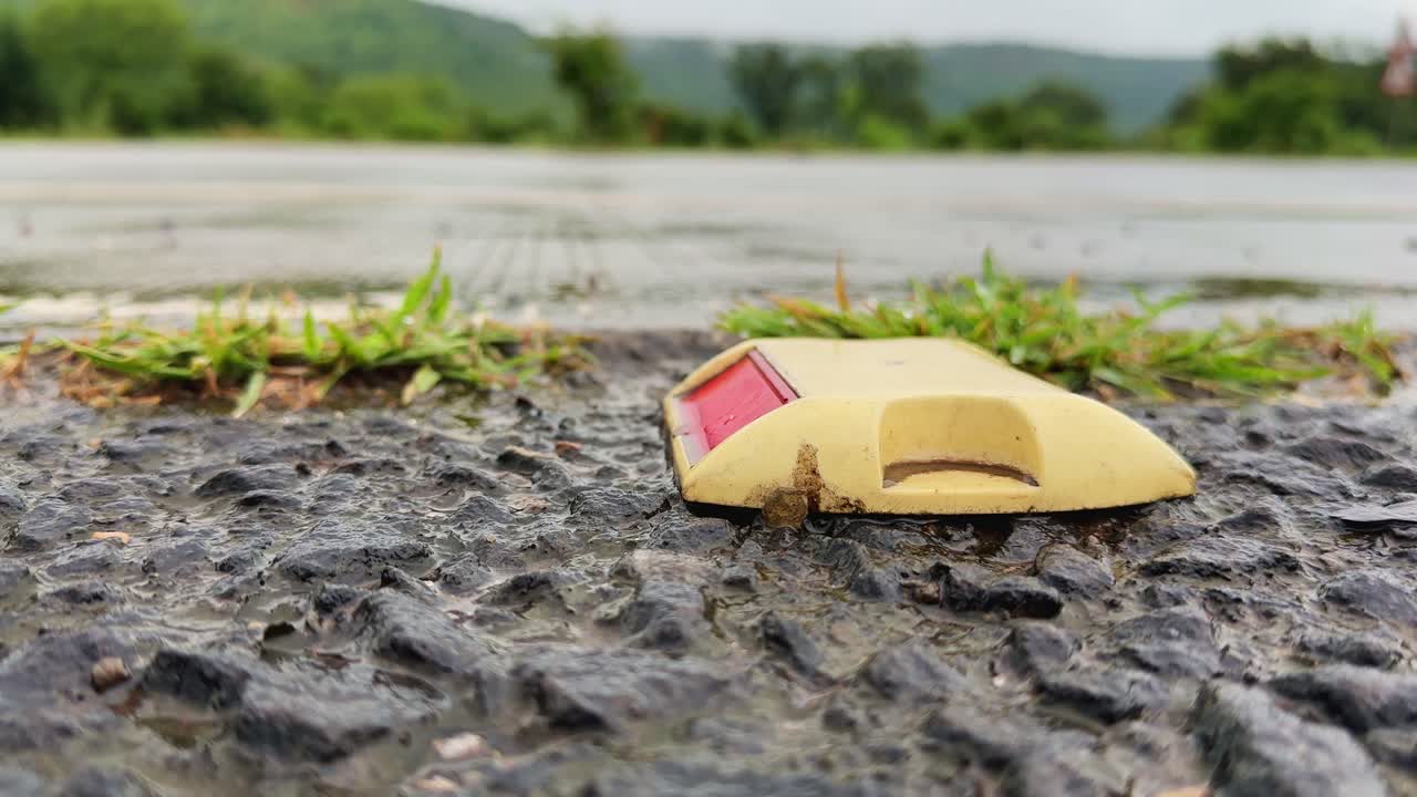 Close-up shot of a yellow road reflector on wet asphalt during rainfall, showing water droplets, texture, and reflective safety detail on a rainy countryside road