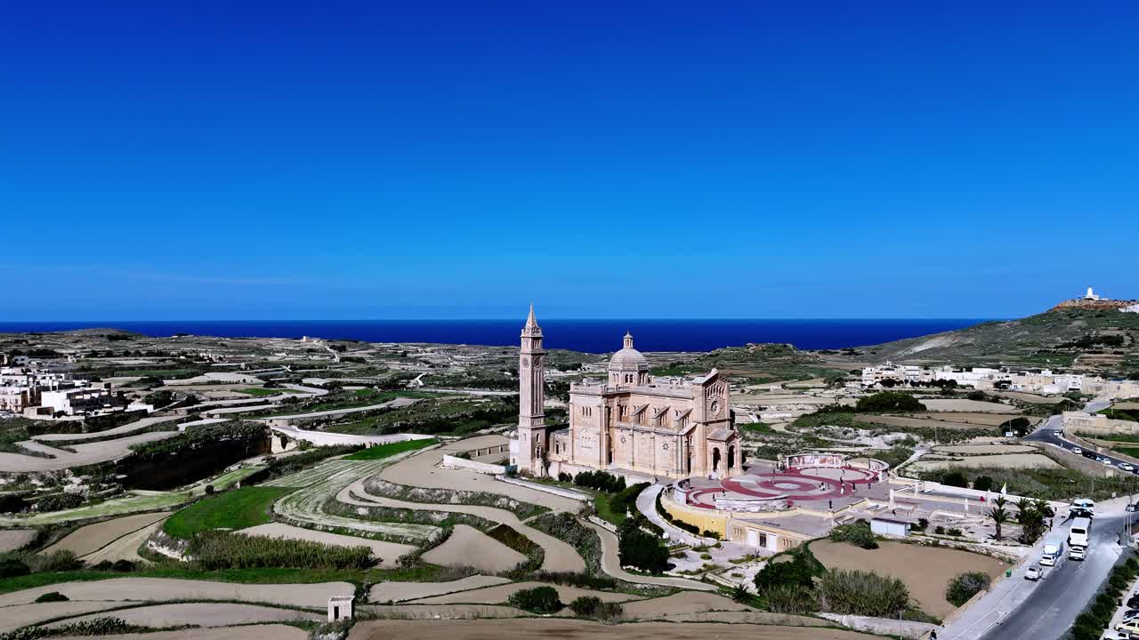 Una maestosa prospettiva aerea rivela il santuario sacro di Ta' Pinu a Gozo, Malta, sotto un cielo azzurro limpido durante la primavera, con un'architettura in pietra calcarea circondata da verdi campi terrazzati e vegetazione mediterranea