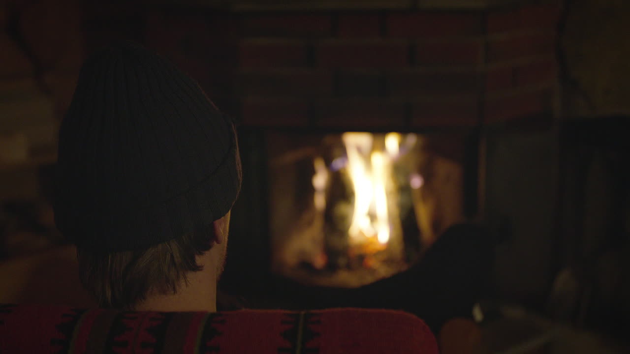 A person in a knit beanie and warm sweater sits near a glowing fireplace, watching the flames, in a dimly lit cozy indoor setting.