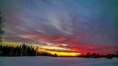 Toma-De-Tiempo-De-La-Puesta-De-Sol-Dorada-Durante-La-Noche-En-Campos-Agrícolas-Cubiertos-De-Nieve-Al-Lado-De-Casas-De-Pueblo-Con-Nubes-Pasando