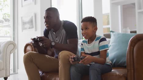 Video-of-happy-african-american-father-and-son-sitting-on-sofa-and-playing-video-games