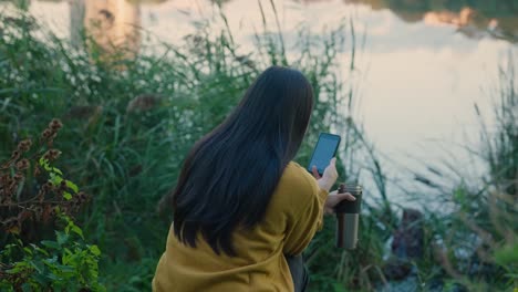 young woman with long hair reads ebook on phone, near lake at sunrise