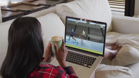 Composite-of-woman-sitting-at-home-holding-coffee-watching-athletics-running-event-on-laptop