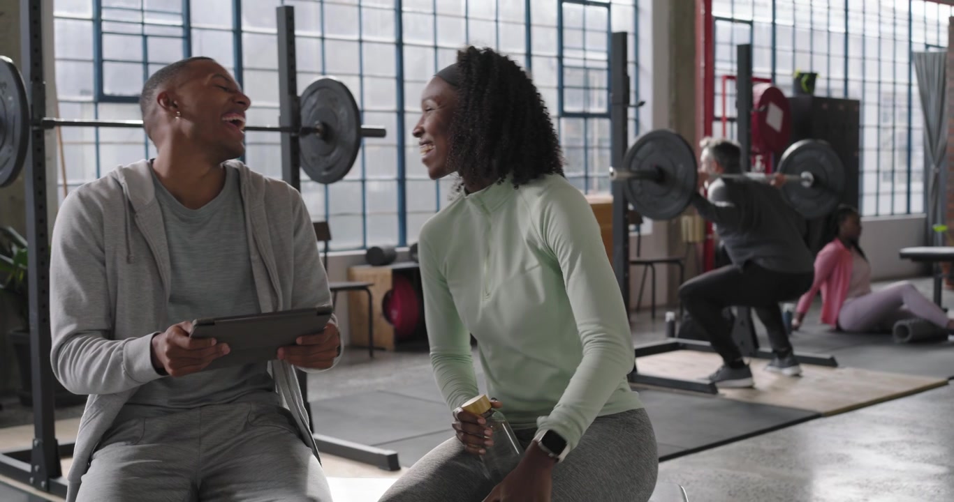 African American friends man tapping tablet, woman leaning in at gym bench reviewing content