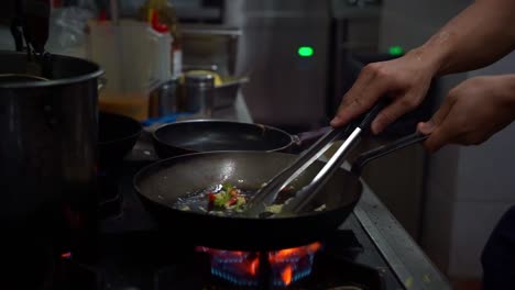 chef stirring, swirling and mixing the ingredients with tongs, sauteing bell peppers with diced garlics in high heat on shallow frying pan