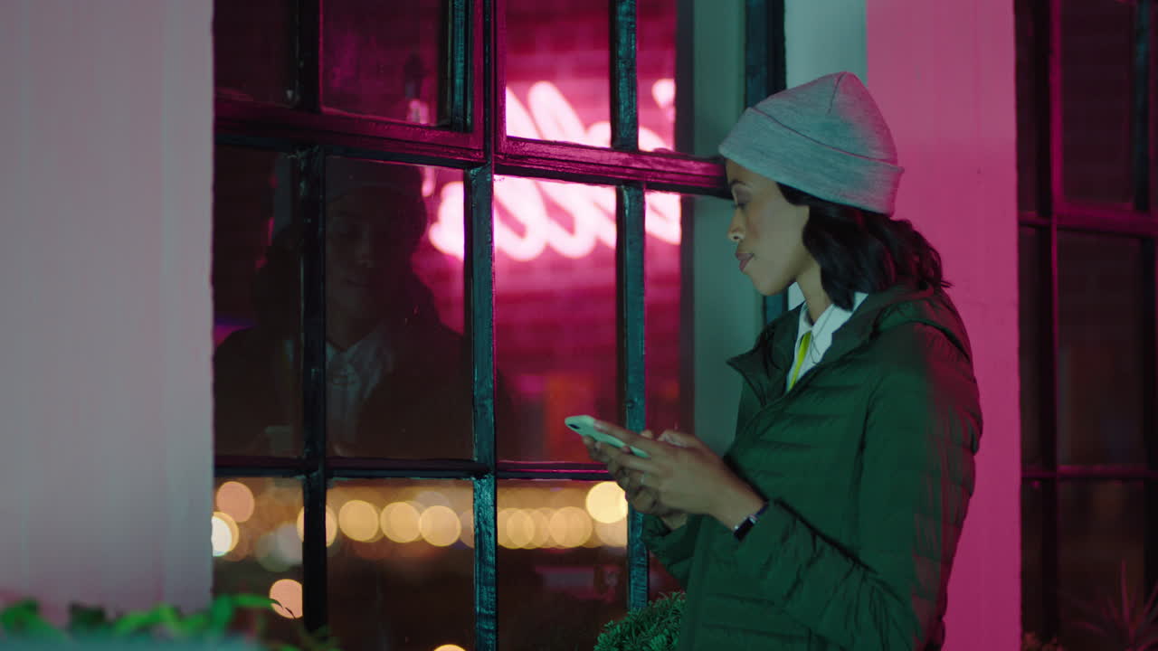 젊은 아프리카계 미국인 사업가 여성이 스마트폰을 사용하여 문자 메시지를 보내고  ⁇ 드폰으로 온라인 소셜 미디어 메시지를 검색하고 창밖을 바라보고 밤에 사무실에서 계획을 세우고 있습니다.