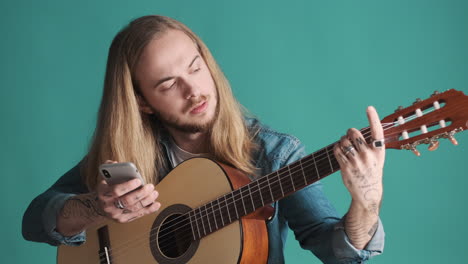 joven caucásico comprobando acordes en su teléfono inteligente y tocando la guitarra en la cámara.