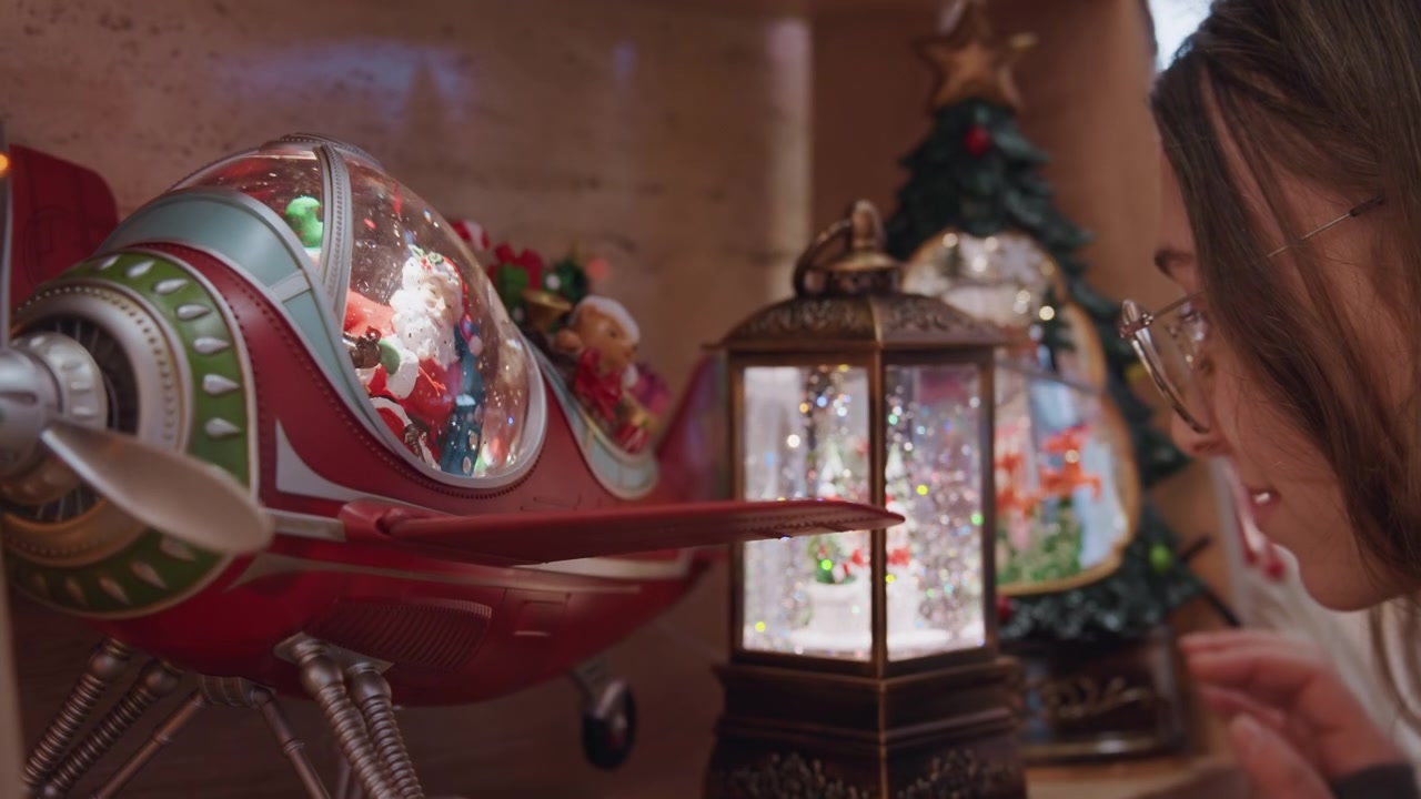 Caucasian woman inspecting vintage christmas ornament in cozy boutique closeup profile with spectacles leaning toward glass snow globe airplane ornament beside lit lantern, warm bokeh lighting