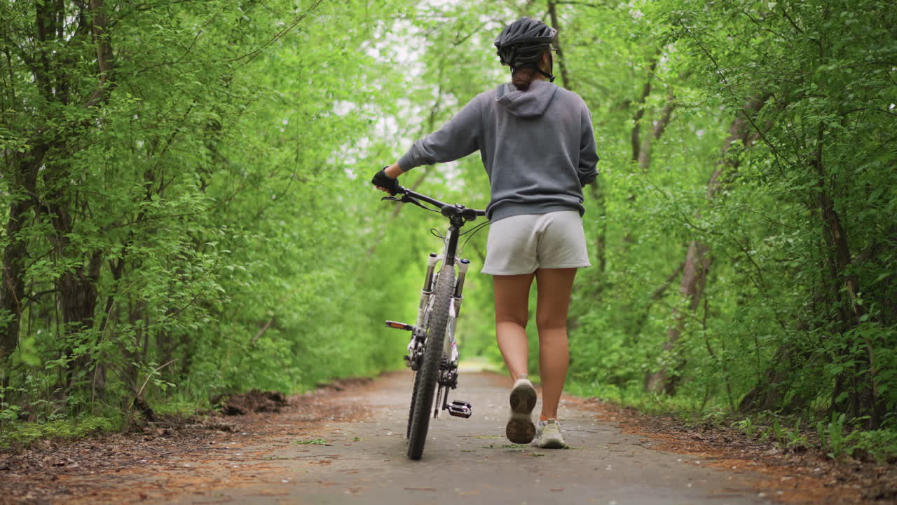 Athlete Biking Along Lush Woodland Trail, Person Cycling Amidst Tall Trees For Health Exercise, Asian Sports Enthusiast Walks Guiding Bicycle On Vibrant Leafy Forest Pathway For Fitness