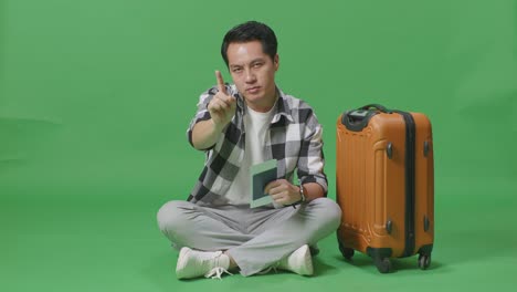 full body of asian male traveler with luggage and passport looking at camera and disapproving with no index finger sign in the green screen background studio