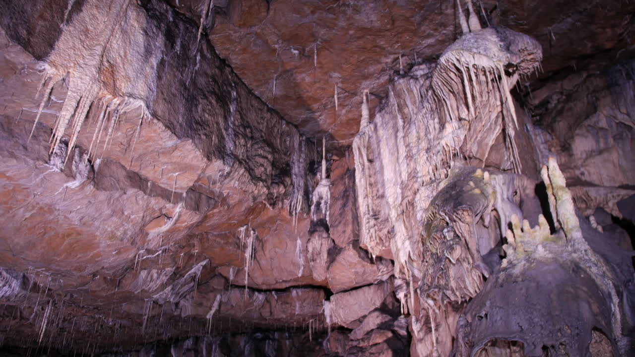 An expansive cave features a ceiling adorned with delicate stalactites and massive rock formations. The rugged textures and formations with intricate geological details