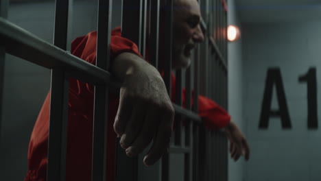 elderly prisoner in orange uniform cries holds his hands on metal bars