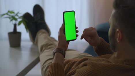 man is relaxing in apartment using mobile phone surfing internet and shopping online green screen on gadget scrolling touch screen