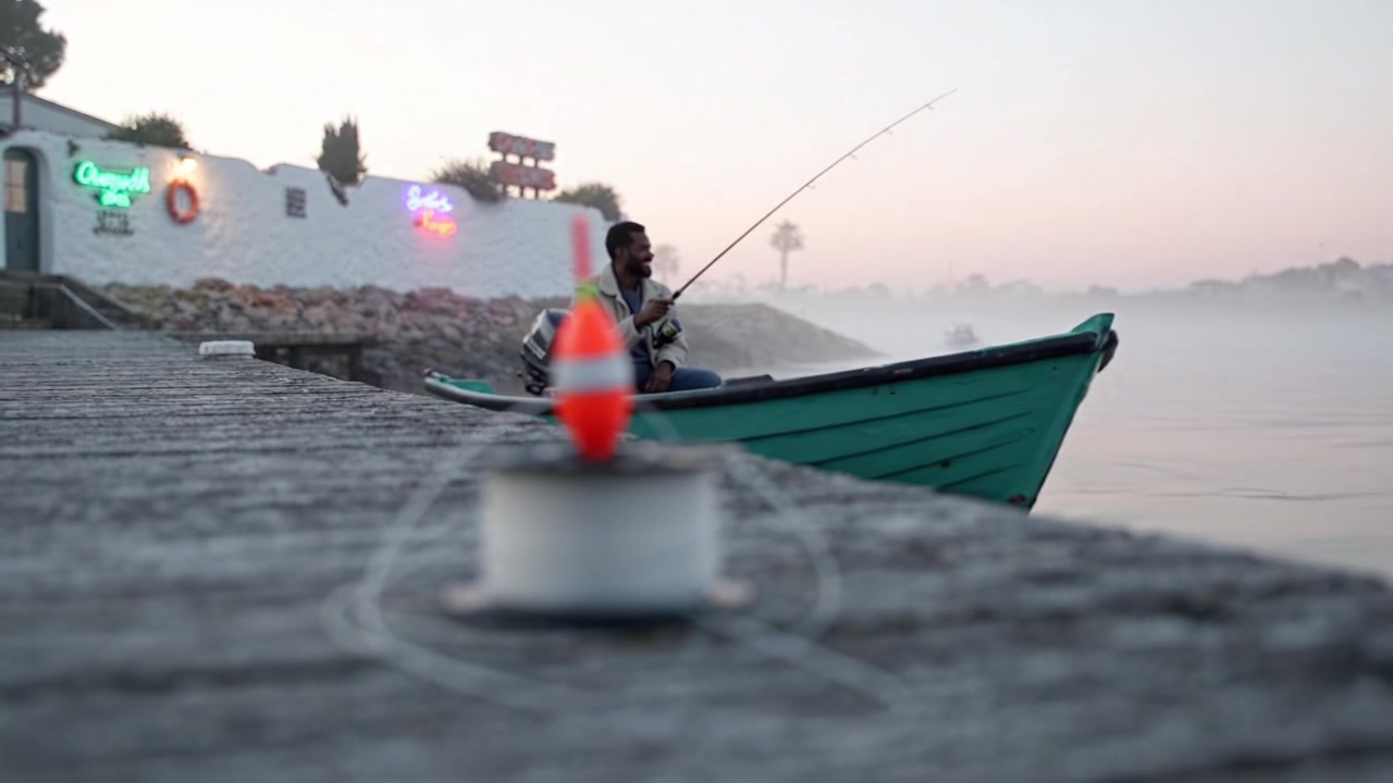 Una tranquilla scena di pesca all'alba, con un galleggiante su una bobina e un pescatore che lancia la sua lenza nelle acque tranquille in mezzo a un paesaggio nebbioso.