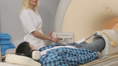 woman doctor makes knee-joint mri scanning. young man patient on automatic table moves outside of closed-type mri machine using noise isolation headphones. modern equipment, coil on the patient's knee