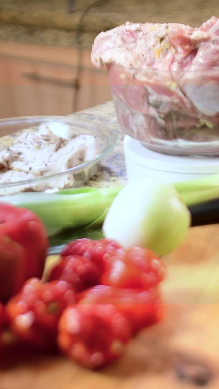 Inquadratura verticale che mostra una varietà di ingredienti freschi su un piano di lavoro in granito, tra cui peperone rosso, cipolla bianca e porro in primo piano. Due ciotole di vetro contengono carne cruda marinata sullo sfondo
