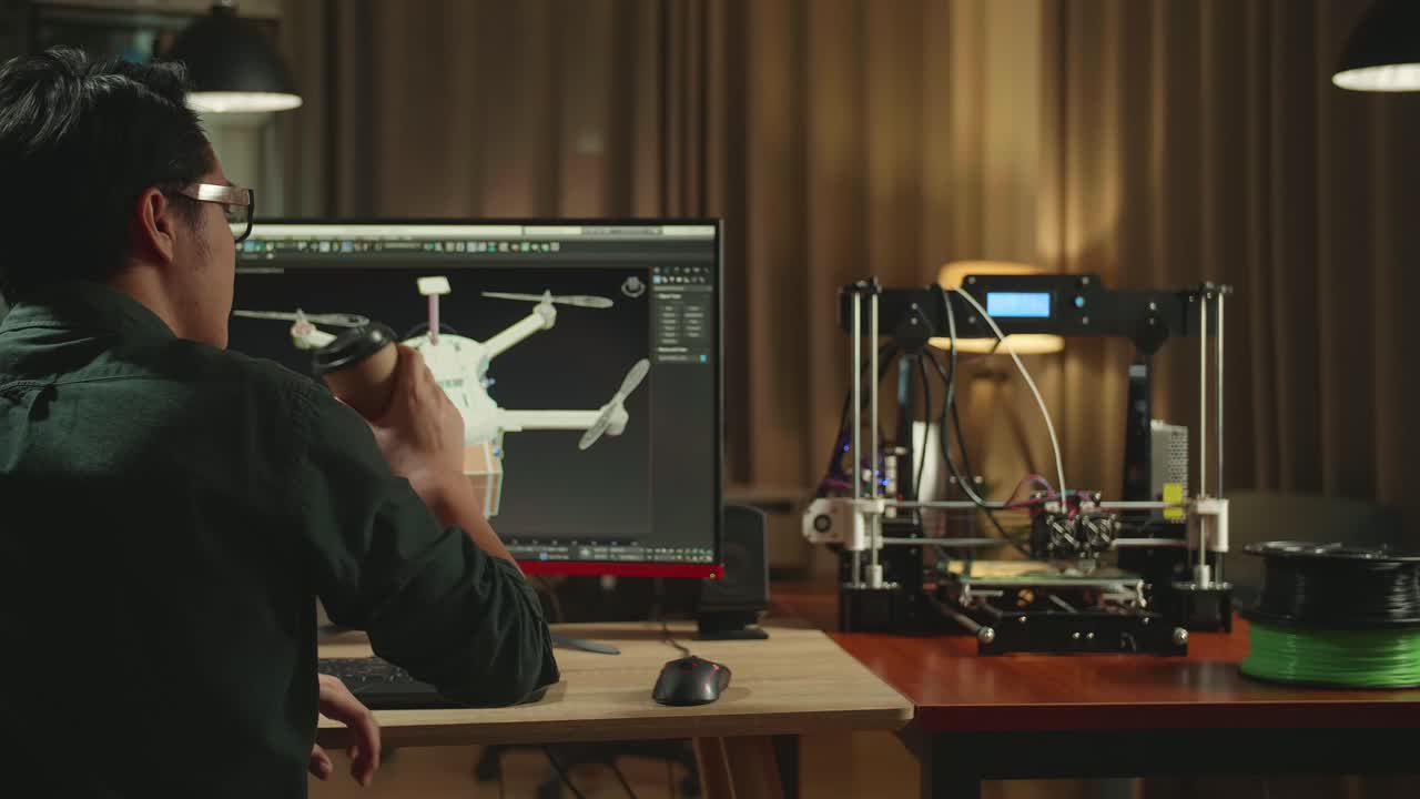Back View Of Asian Engineer Drinking Coffee While Work On Personal Computer And 3D Printer, Screen Shows Cad Software With 3D Prototype Of Drone Delivery