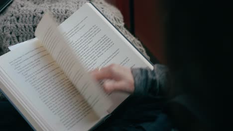 la mano de una mujer volteando una página mientras lee un libro, en casa con una manta en