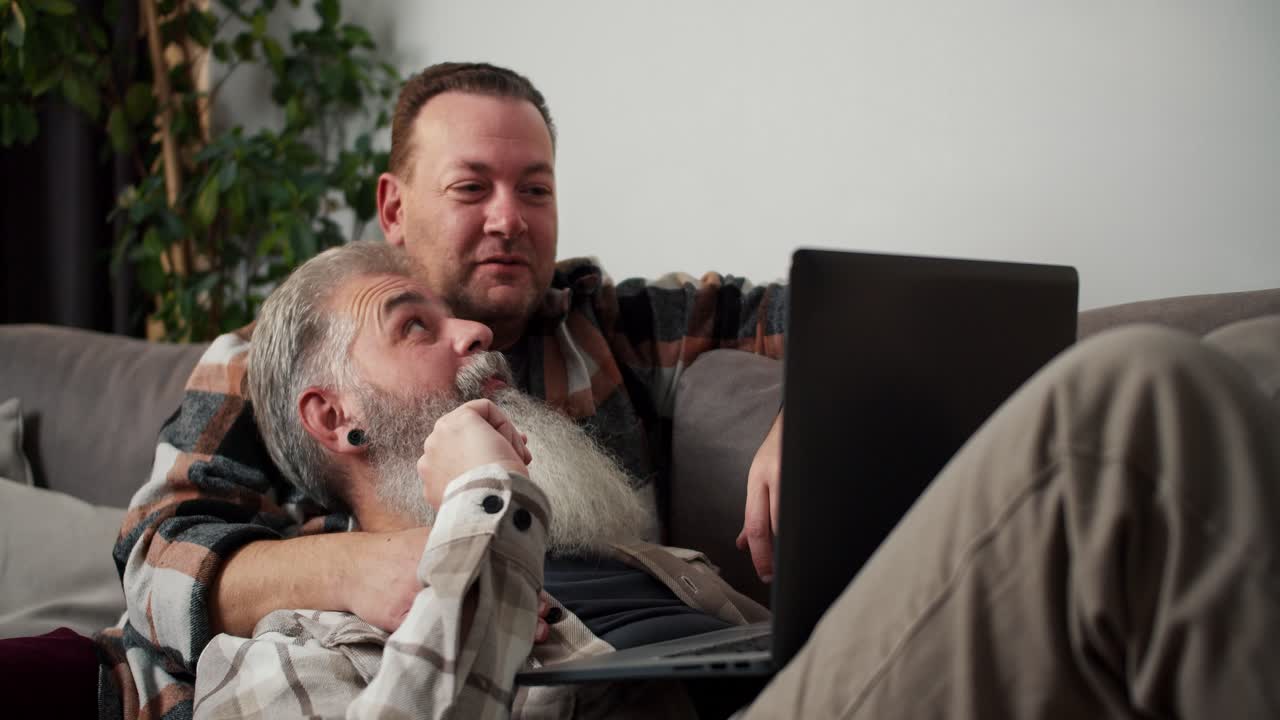 un hombre moreno feliz en una camisa a cuadros mira una computadora portátil gris que está sosteniendo. su novio un hombre de mediana edad con cabello gris y una barba en una camiseta a cuadros yace en sus brazos en el sofá en un apartamento moderno