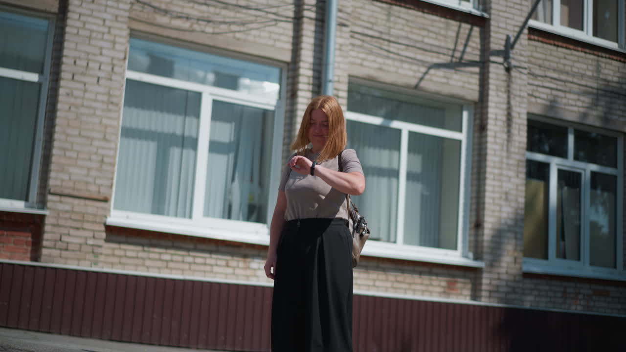 Round view on female individual standing under strong summer sunlight near building, looking around and checking time with fitness bracelet, appearing exhausted and thoughtful