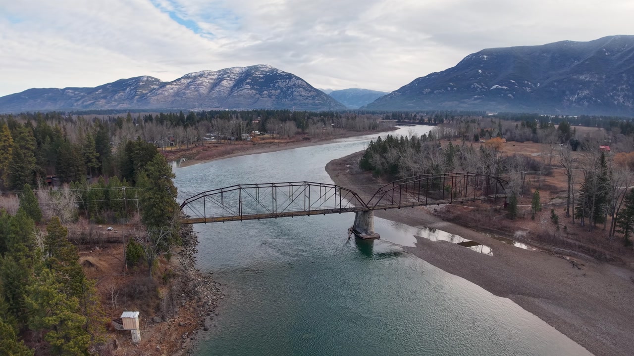 Cinematic aerial shot of a river winding past forested banks, with a narrow bridge connecting the shores and mountains looming in the background under soft clouds.