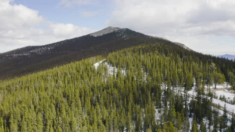 low aerial reveal forward of sunny and snow covered back roads in the colorado mountains surrounded by bright green pine tree forests with blue skies