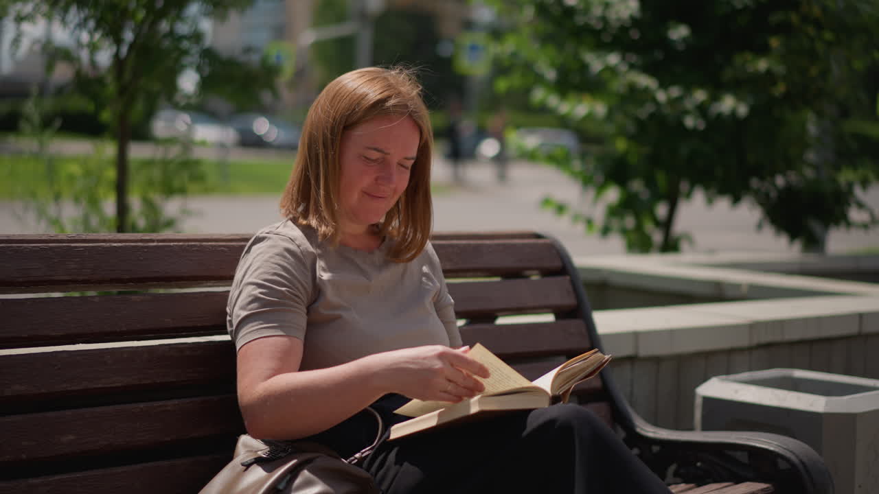 Étudiant assis sur un banc en bois, étudiant un livre et tournant une page sous une lumière vive, expression calme et esprit concentré entouré d’arbres verts et d’un arrière-plan doux