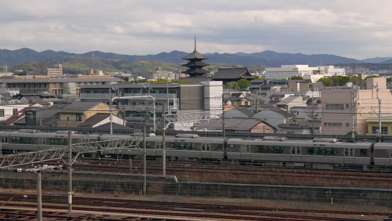 日本の京都鉄道博物館から見た線路の外観を捉えた静止画。地元列車が通り過ぎ、遠くには象徴的な東寺の五重塔が背景に見える。