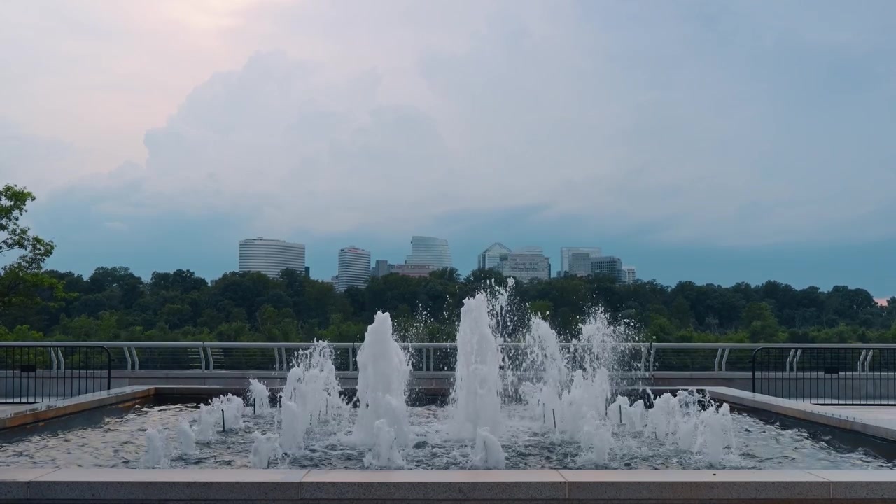 Fuente de agua con el horizonte de Rosslyn de fondo, Washington D.C. Chorros de agua rociando sobre una pila de piedra, barandilla de metal, árboles y cielo nublado al anochecer