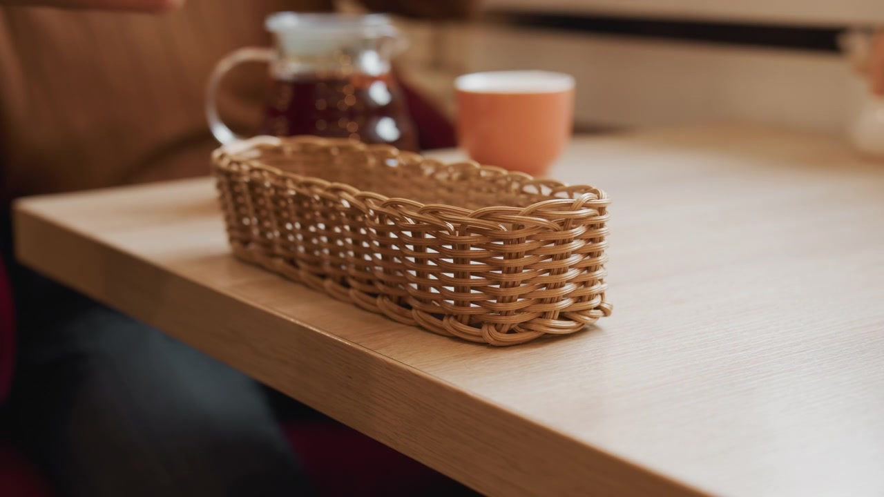 Primo piano delle mani di una persona che posiziona e sistema un cesto di vimini intrecciato su un tavolo di legno chiaro. Una caffettiera di vetro con liquido scuro e una tazza arancione sono visibili sullo sfondo, suggerendo un ambiente interno.