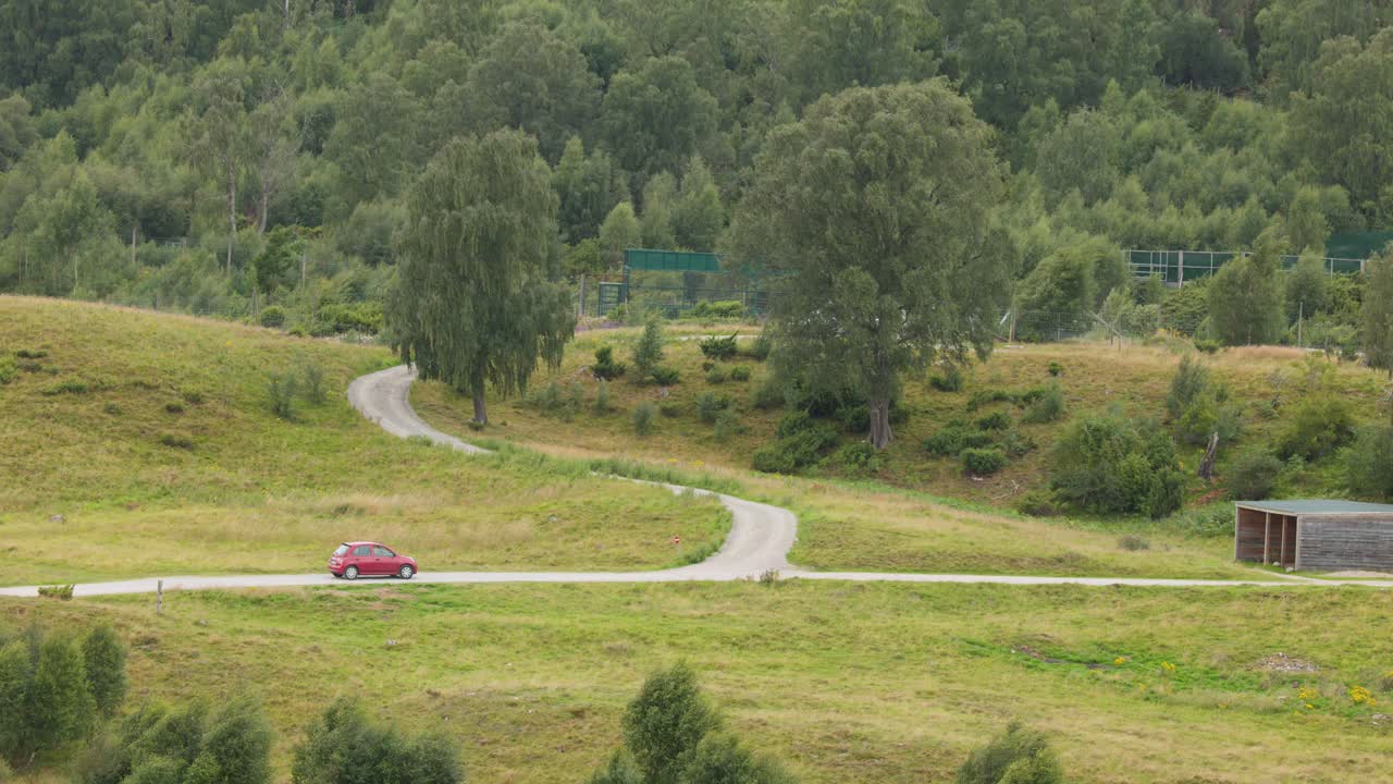 Red car travels along curving rural road through grassy highlands, daylight, wide static shot