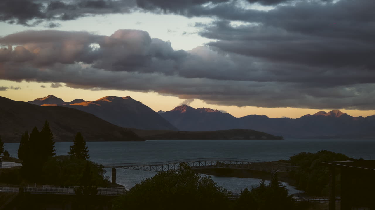 Timelapse image of Lake Tekapo during sunset, showcasing glowing clouds, calm waters, and distant snowy mountain peaks