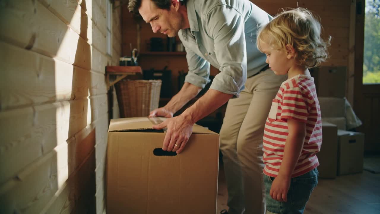 Parent bringing moving box into wooden room, lowering and opening flaps while child watching