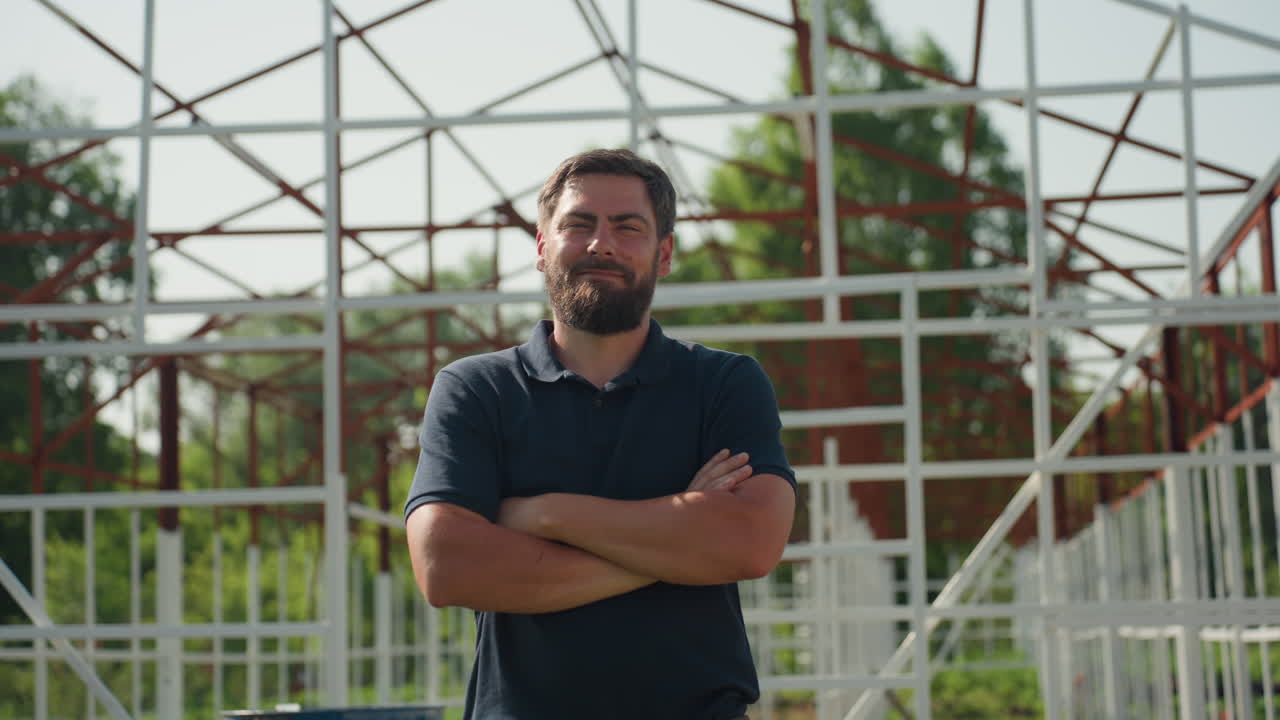 Granjero girando con las manos cruzadas frente a un edificio con estructura metálica en un día soleado, con expresión tranquila y vegetación alrededor, inspeccionando la propiedad y el progreso de la estructura durante un proyecto de construcción rural.