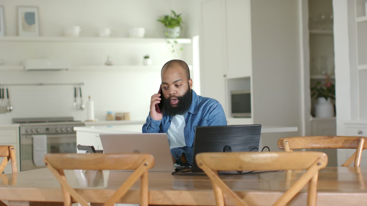 African American man talking on smartphone, typing between two laptops for work at kitchen table