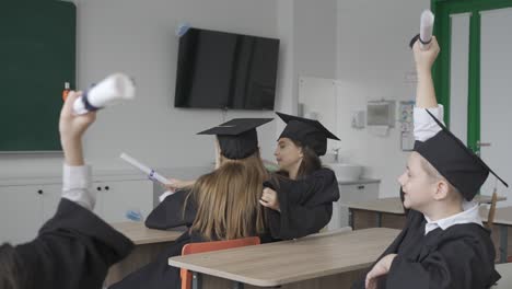 two happy students hugging in class. they are wearing gowns and mortarboards.