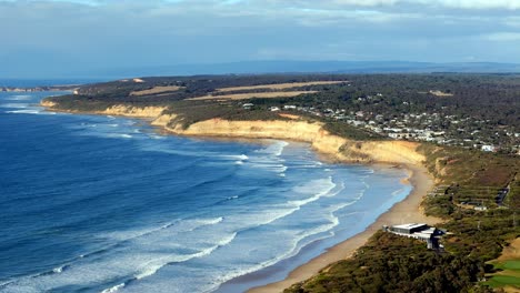 Coastal-cliffs-tower-over-a-sweeping-sandy-beach,-Waves-gently-breaking-along-the-shoreline-with-a-quaint-town-in-the-distance