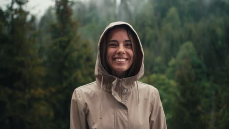 Portrait-of-a-blonde-girl-in-a-jacket-with-a-hood-posing-and-smiling-against-the-backdrop-of-a-green-mountain-forest